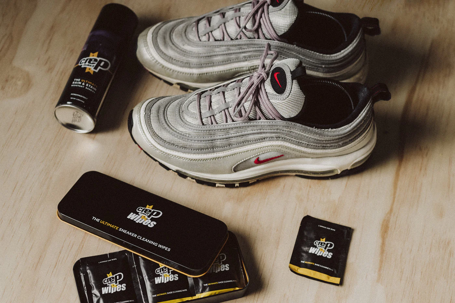 A pair of grey Nike Air Max 97 sneakers placed on a wooden surface, surrounded by Crep Protect cleaning products, including a spray can, a tin of sneaker wipes, and individual wipe packets.