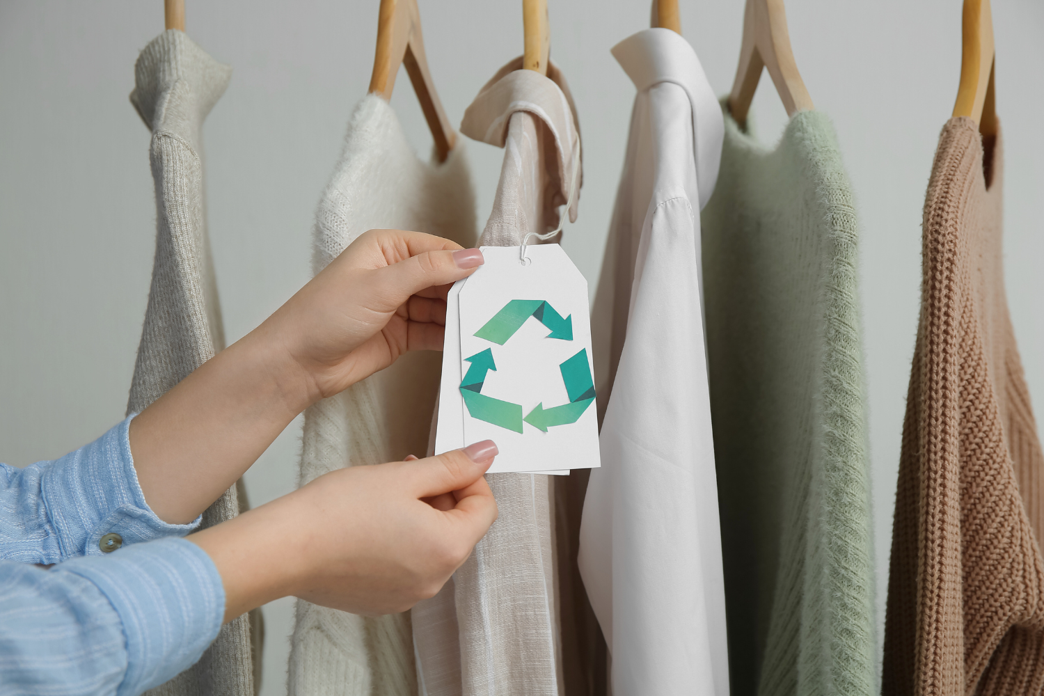 &ldquo;Hands holding a clothing tag with a green recycling symbol in front of a row of neutral-toned garments on wooden hangers, including sweaters, a blouse, and knitwear.
