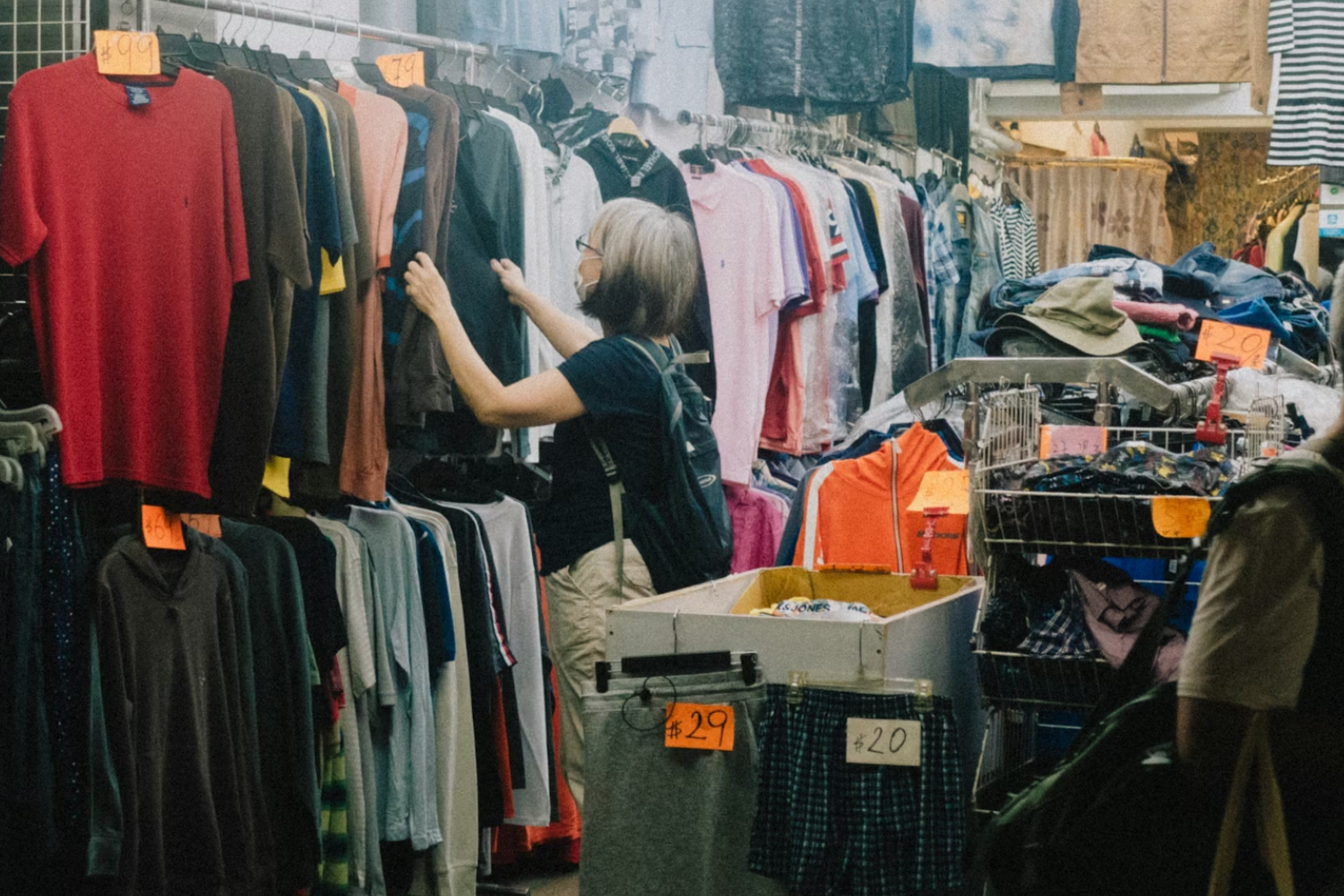 Person browsing secondhand clothing at a market stall with racks of shirts and visible price tags.