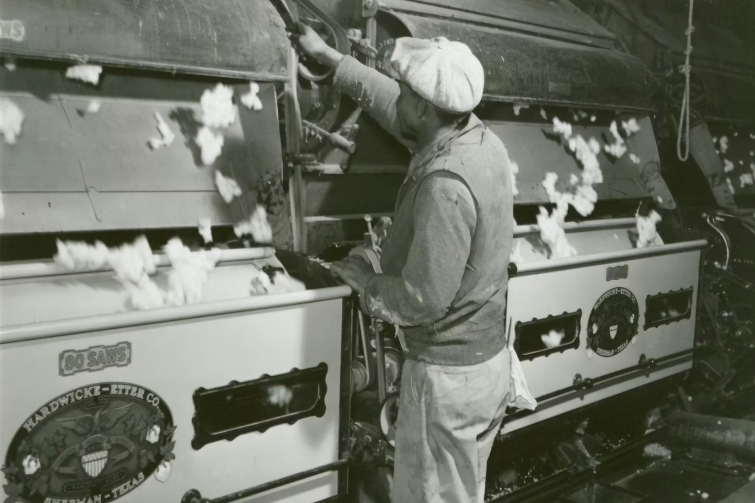 Black-and-white photo of a factory worker operating textile machinery, with loose fibers floating in the air.