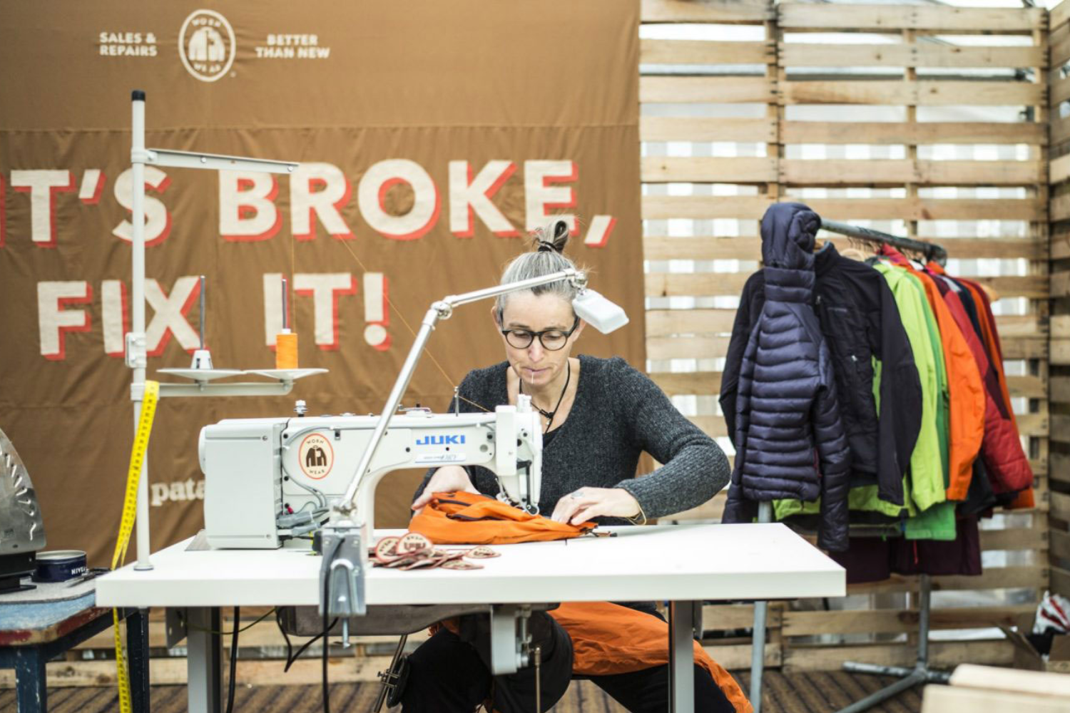 Person repairing an outdoor jacket at a sewing machine, with a sign reading &ldquo;It&rsquo;s broke, fix it&rdquo; in the background.